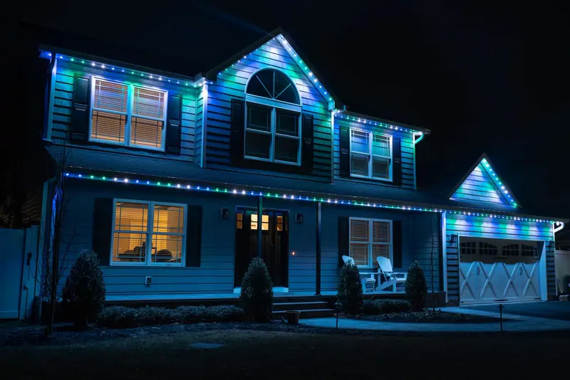 Two-story home glowing blue and teal, with lights tracing the roofline and gables