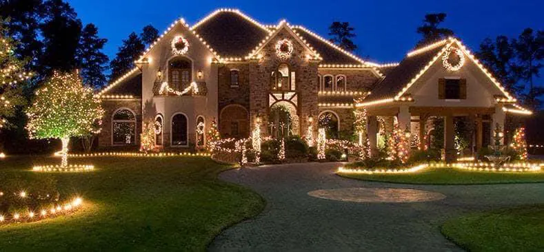 Large stone home with warm white lights, wreaths and lit trees in the front garden